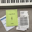 Fotografie zachycuje část klávesnice s bílými a černými klávesami a přiloženou dokumentaci. V popředí je zelený sešit s nápisy „Song BooK“ a logem „C…