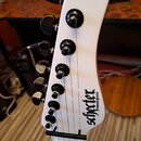 Bílá hlava elektrické kytary s šesti černými ladičkami. Na povrchu je černý nápis schecter a pod ním menší text Diamond Series. Nástroj vykazuje znám…