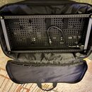 Černý efektový pedalboard s perforovanou deskou a bočními lištami, umístěný v otevřeném černém polstrovaném pouzdře. Na desce je položen černý napáje…