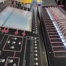 Digitální mixovací pult s černým povrchem a červenými detaily. Na levé části je dotykový displej, řada červených tlačítek a ovládací prvky s nápisy s…