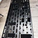 Černý kovový pedalboard s mřížkovým dnem. Na povrchu jsou nalepené různé útržky černého suchého zapojení (velcro). Povrch vykazuje běžné známky použí…