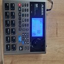 Šedý bicí automat s černými ovládacími tlačítky a modrým displejem. Na horním panelu jsou nápisy Alesis, SR18 a DRUM MACHINE WITH DYNAMIC ARTICULATIO…