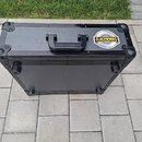 Černý transportní kufr s rukojetí a kovovými hranami. Na přední straně je nalepená kulatá nálepka s nápisem FLACHWORKS. Povrch vykazuje lehké známky…