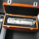 Stříbrný kondenzátorový mikrofon v dřevěném kufru s černým polstrováním. Na těle mikrofonu je viditelné kulaté logo se písmenem „S“. Kufřík má na pře…