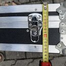 Detailní pohled na část černého transportního kufru s hliníkovými hranami a kovovými rohy. Uprostřed je umístěn chromovaný zámek. Vedle něj je přilož…