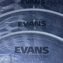Tři průhledné bubny značky EVANS s označením G2. Nápisy jsou provedeny černým tiskem. Povrch materiálů vykazuje drobné škrábance a opotřebení.