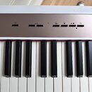 Detailní pohled na klávesnici digitálního piana s bílými a černými klávesami. Nad klávesami je umístěn ovládací panel s několika tlačítky a symboly,…