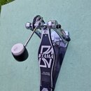Černý bicí pedál s kovovými mechanismy a bílým plátěným špičákem. Na stupni je bílý nápis TAMA a pod ním text THE LEGEND IN INNOVATION. Povrch stupně…