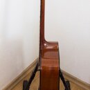 Fotografie zachycuje akustickou kytaru z profilu, umístěnou na černém stojanu. Nástroj má hnědý lakovaný povrch s viditnou strukturou dřeva. Na hlavě…