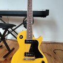 Žlutá elektrická kytara typu Les Paul s černým pickguardem a černými knoflíky. Na hlavě hmatníku je bílý nápis "Tokai". Nástroj je umístěn na černém…