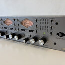 Stříbrné rackové audio zařízení s nápisem UNIVERSAL AUDIO a doprovodným logem. Přední panel obsahuje tři analogové VU metry, několik otočných knoflík…