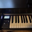 Digitální piano v černém provedení s bílými a černými klávesami. Na ovládacím panelu je displej s nápisem SK Concertgrand a řada tlačítek s popisky j…