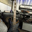 Elektrická basová kytara s tmavým, žilnatým povrchem těla a hnědými okraji. Na hlavě nástroje je viditelný nápis Marcus Miller. V pozadí jsou umístěn…