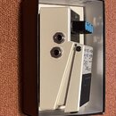 Bíle provedené elektronické zařízení v kartonové krabici. Na boku jsou dva vstupy typu jack a tři modré ovládací knoflíky. Je zde čitelně napsán nápi…