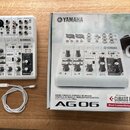 Bílá mixovací konzole Yamaha AG06 s černými ovládacími prvky a připojeným bílým USB kabelem. Vedle nástroje je umístěna originální kartonová krabice…