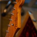 Fotografie zachycuje hlavu elektrické kytary z přírodního dřeva. Na povrchu je viditelné logo Fender a nápis STRATOCASTER, pod kterým jsou menší, čás…