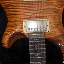 Detail elektrické kytary s hnutým javorovým topem v hnědé barvě. Viditelný je jeden humbucker s krémovým rámečkem a hnedbroudy. Na hmatníku jsou vykl…