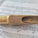 Zlatavý kovový mouthpiece s otvorem pro trsku, položený na notovém papíře. Povrch vykazuje známky opotřebení, drobné škrábance a oxidaci. Na nástroji…