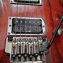 Detail elektrické kytary s červeným lakovaným povrchem s viditelnou strukturou dřeva. Na mostku je vyryto logo Ibanez a na snímači v pozici kobylníku…
