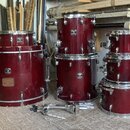 Sada červených bubnů s chromovaným kováním. Na korusech jsou viditelné loga „GRETSCH Custom Style“. Povrch největšího bubnu vykazuje v dolní části od…
