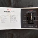 Fotografie znázorňuje otevřené balení malého černého sluchátkového zesilovače. Na zařízení je bílý nápis Fender MUSTANG MICRO. Krabice obsahuje sezna…
