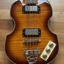 Elektrická basová kytara s tvarem připomínajícím housle a povrchem v barvě sunburst. Nástroj má dva snímače s kovovými kryty, chromovaný most a tři z…