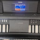 Černý digitální klavír značky YAMAHA s modrým displejem, na kterém jsou viditelné texty jako „Bright Trumpet“ a „DarkMoon“. Před klávesami je umístěn…