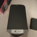 Tmavě šedý elektronický přístroj s obdélníkovým tvarem a žebrovaným chladičem v horní části. Na předním světlejším panelu je umístěno tlačítko a nápi…
