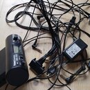 Na dřevěné podlaze leží černý elektronický přístroj značky Roland s nápisem Roland VH-10, napájecí adaptér a několik propojovacích kabelů. Přístroj m…