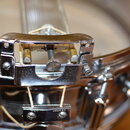 Detailní záběr na chromovaný mechanismus snare drumu. Na kovovém kusu v dolní části je vyryt nečitelný nápis. Povrch chromu vykazuje drobné škrábance…
