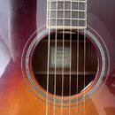 Detail akustické kytary s červeně-oranžovým přechodem barvy (sunburst). Uvnitř otvoru těla je vidět bílá štítek s nápisy YAMAHA, FG-TA a transAcousti…