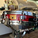 Červený snare drum s chromovanými obručmi a ladicími šrouby. Na korpusu je zlatý nápis TAMA a štítek s textem SNARE DRUM, LEGAL No. 200, MADE IN JAPA…