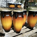 Sada perkusních nástrojů v oranžovo-hnědém přechodovém odstínu s bílými membránami. Na přední straně čtyř velkých bubnů jsou viditelné loga „Headline…