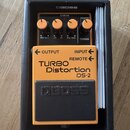 Kytarový efekt v oranžovém provedení s černým pedálem, umístěný v černé krabici značky BOSS. Na přístroji jsou nápisy TURBO Distortion DS-2, OUTPUT,…