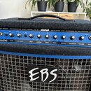 Modrý basový kombo zesilovač v šedém látkovém potahu s kovovou mřížkou. Na ovládacím panelu jsou nápisy EBS DROME PROFESSIONAL BASS COMBO 150 W a DEV…