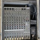 Šedý mixážní pult značky SAMSON, model MX124, umístěný v černé polstrované přepravní skříni. Zařízení má více vstupních kanálů s posuvníky, otočnými…