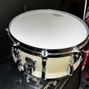Bílá buben (pravděpodobně snare) s chromovaným obručováním a ladicími šrouby. Na bláně je viditelné logo REMO. Korpus má krémovou barvu a na mechanis…