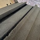 Detailní záběr na černou pěnovou vložku s vyfrézovanými drážkami. Materiál má hrubou, pórovitou strukturu. V pozadí je vidět část světlého dřevěného…