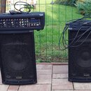 Dva černé reproduktory značky ALESIS s kovovými mřížkami. Na levém reproduktoru je umístěný černý mixážní pult s nápisy ALESIS a PAM. Na pultu jsou v…