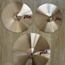 Tři mosazné činely s viditelným broušením povrchu položené na dřevěné podlaze. Všechny tři mají červenobílé logo pAiste pst 1. Činely mají nápisy 14"…