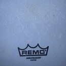Fotografie zobrazuje bílou, texturovanou plochu bubnové skins. Uprostřed je černé logo s nápisem REMO pod stylizedovanou korunou a pod ním text AMBAS…