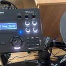 Černý elektronický bicí modul s nápisy Roland a V-Drums. Na displeji je vidět text Deep Studio a Harp and Close. Dole na zařízení je uveden model DRU…