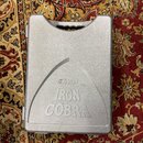 Šedý plastový kufřík s úchytem v horní části. Na přední straně je vyražen nápis TAMA a pod ním výrazný text IRON COBRA. Povrch plastu je matní a vyka…