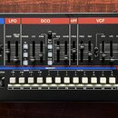 Černý syntetizátor s ovládacími prvky v horní části rozdělenými do barevných sekcí: KEYBOARD, ARPEGGIO, LFO, DCO, HPF, VCF, VCA a ENV. Na přední stra…