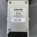 Fotografie zachycuje spodní stranu stříbrného kytarového pedálu. Na bílém štítku je uvedeno logo NUX, nápis AC ADAPTER: DC 9V a symboly CE a koš. Dál…