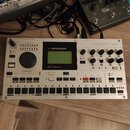 Bílý elektronický bubnový stroj s nápisy Elektron a MACHINEDRUM. Zařízení obsahuje displej, několik otočných knoflíků, tlačítka pro výběr zvuků a sek…