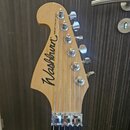 Fotografie zachycuje hlavu elektrické kytary z přírodního světlého dřeva. Na ní je černé logo Washburn® a nápis NUNO BETTENCOURT Model. Jsou vidět še…