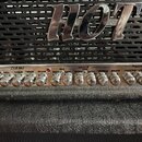 Detail kytarového zesilovače s kovovým nápisem HOT na černé mřížce. Pod ním je ovládací panel s řadou stříbrných knoflíků rozdělený na sekce CLEAN, D…