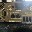 Zadní strana šedého elektronického zařízení s kovovým povrchem. Je zde vidět ventilátor s ochrannou mřížkou, vstup AC LINE IN a dva výstupy SPEAKERS.…