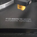 Detailní záběr na černý gramofon s nápisem F·G·SERVO MOTOR AUTOMATIC TURNTABLE. Na raménku je nasazen žlutý hloubkový systém s logem Audio-Technica.…